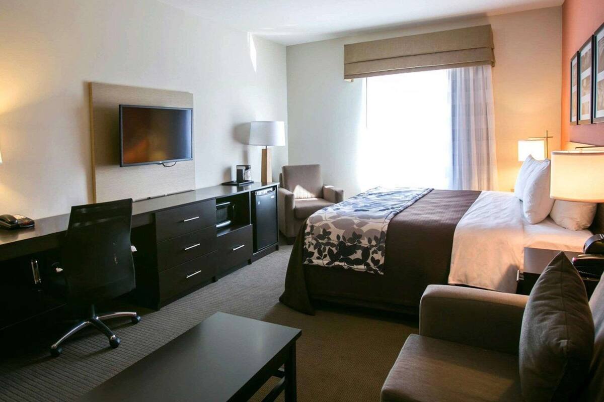 A hotel room with a bed, a desk with a chair, a television, and a window with curtains.
