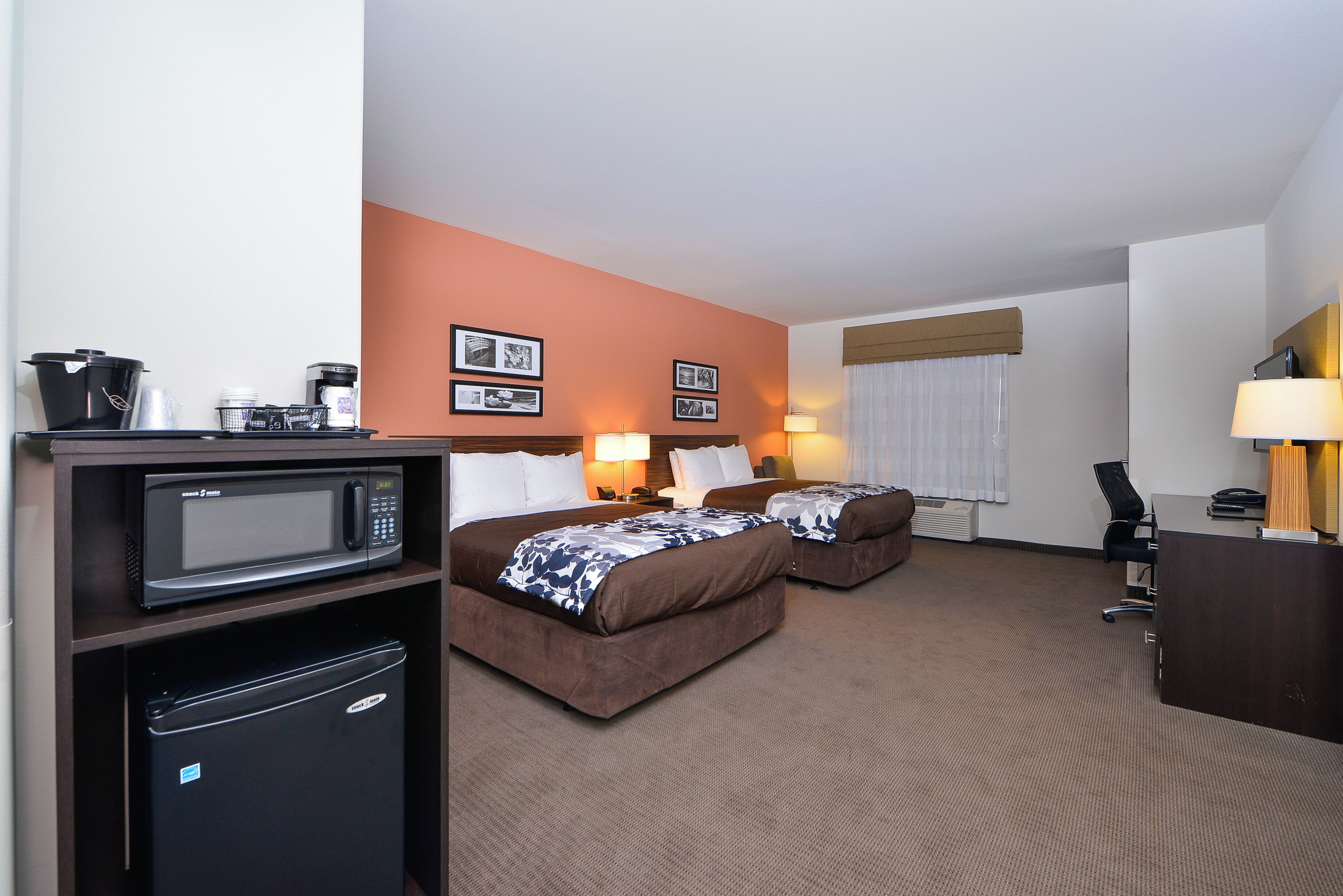 A hotel room with two beds, a microwave, a refrigerator, a desk, and a window with curtains.