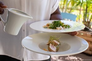 A person holding two white plates with food, one with a green garnish and the other with a piece of meat, in an outdoor setting with greenery in the background. - Diamant les Bains Résidence Hôtelière (Le Diamant)