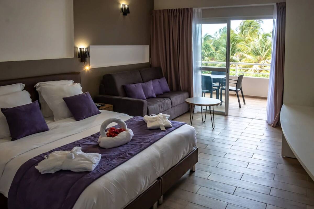 Ein Hotelzimmer mit einem großen Bett, einer Sitzecke, einem kleinen Tisch und einem Balkon mit Blick auf Palmen.
