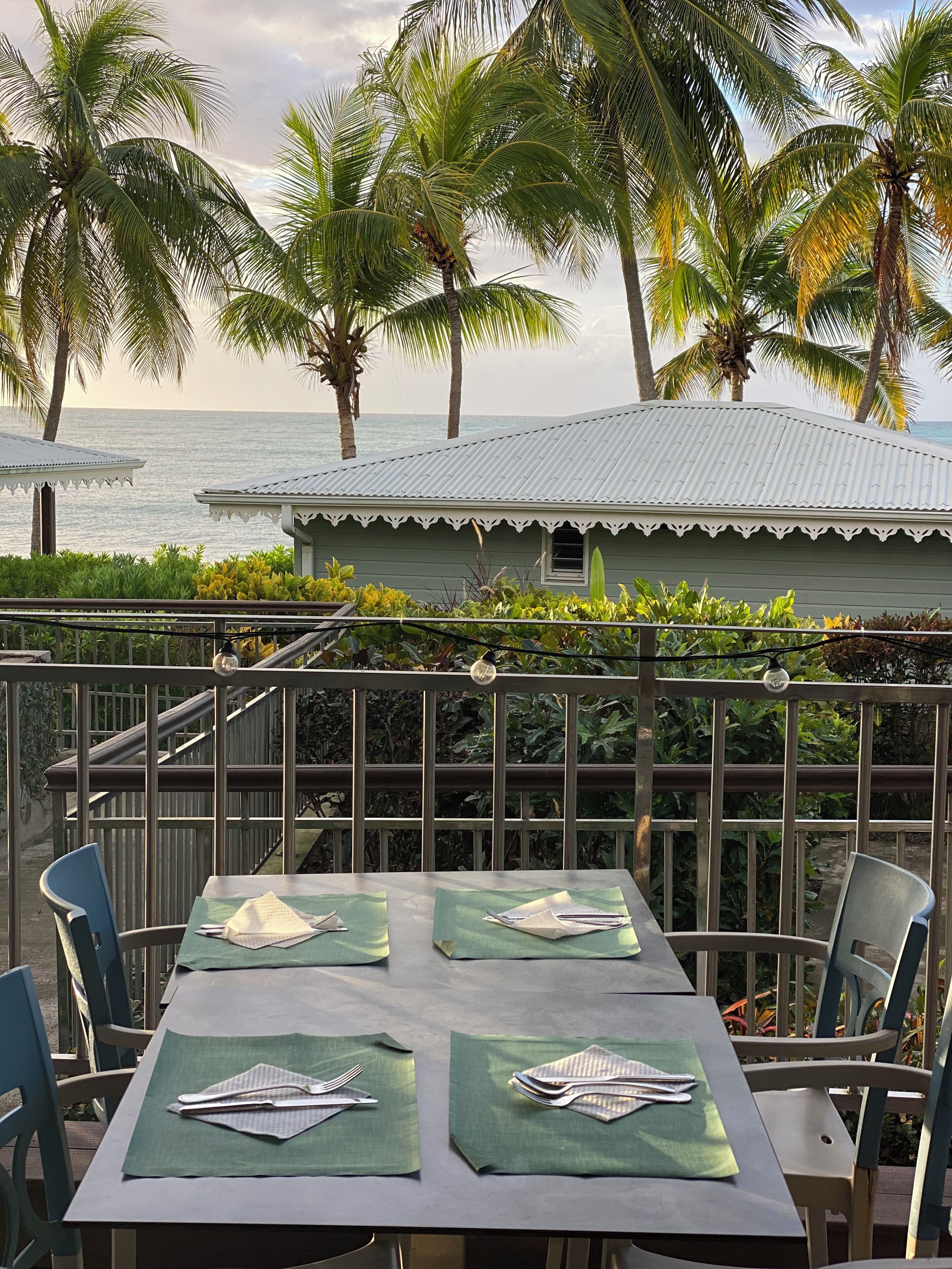 A dining area with tables set for two, overlooking a beach with palm trees and a building with a corrugated metal roof.