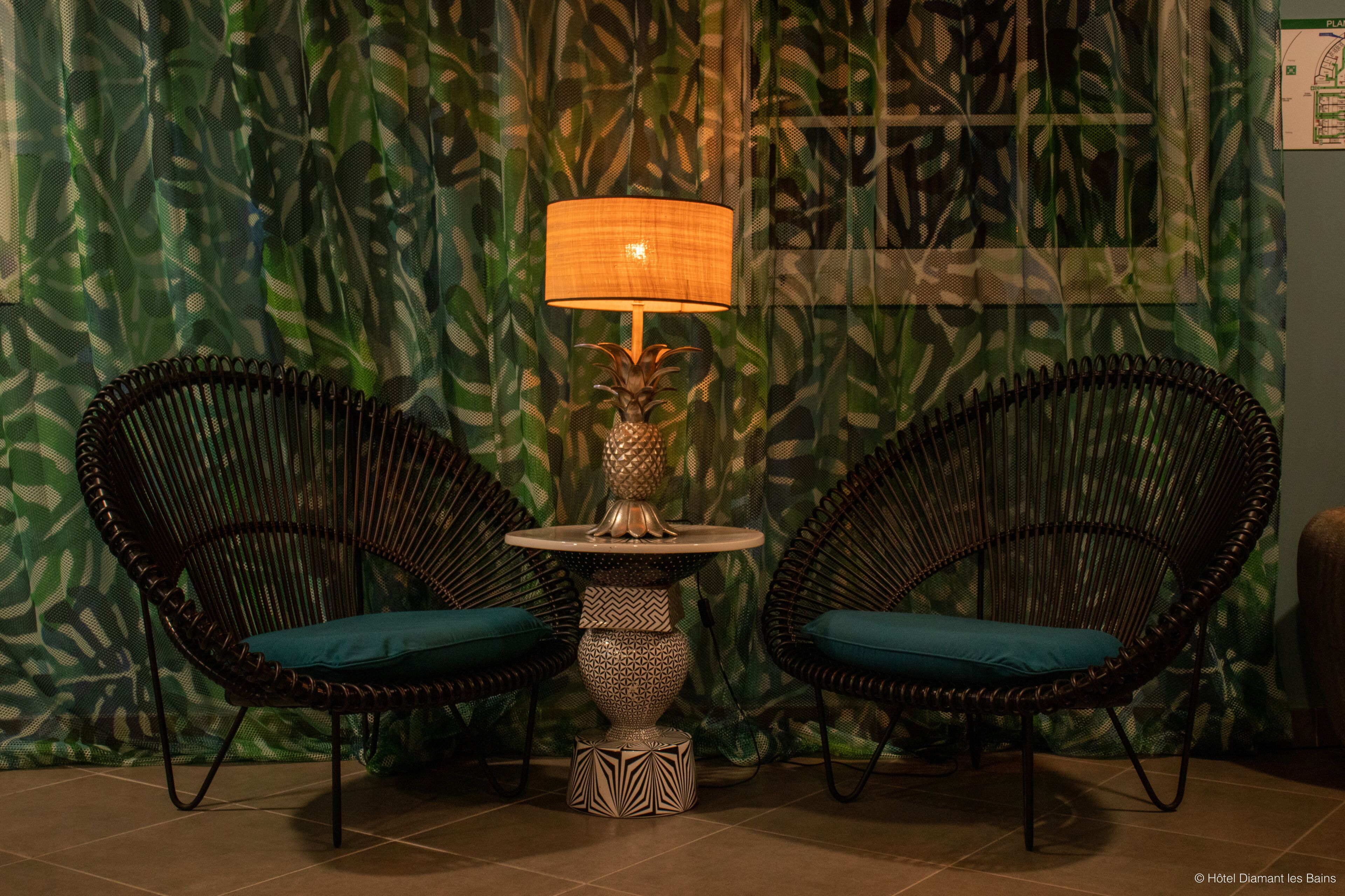 A room with two wicker chairs with teal cushions, a small round table with a decorative lamp, and a patterned wall.