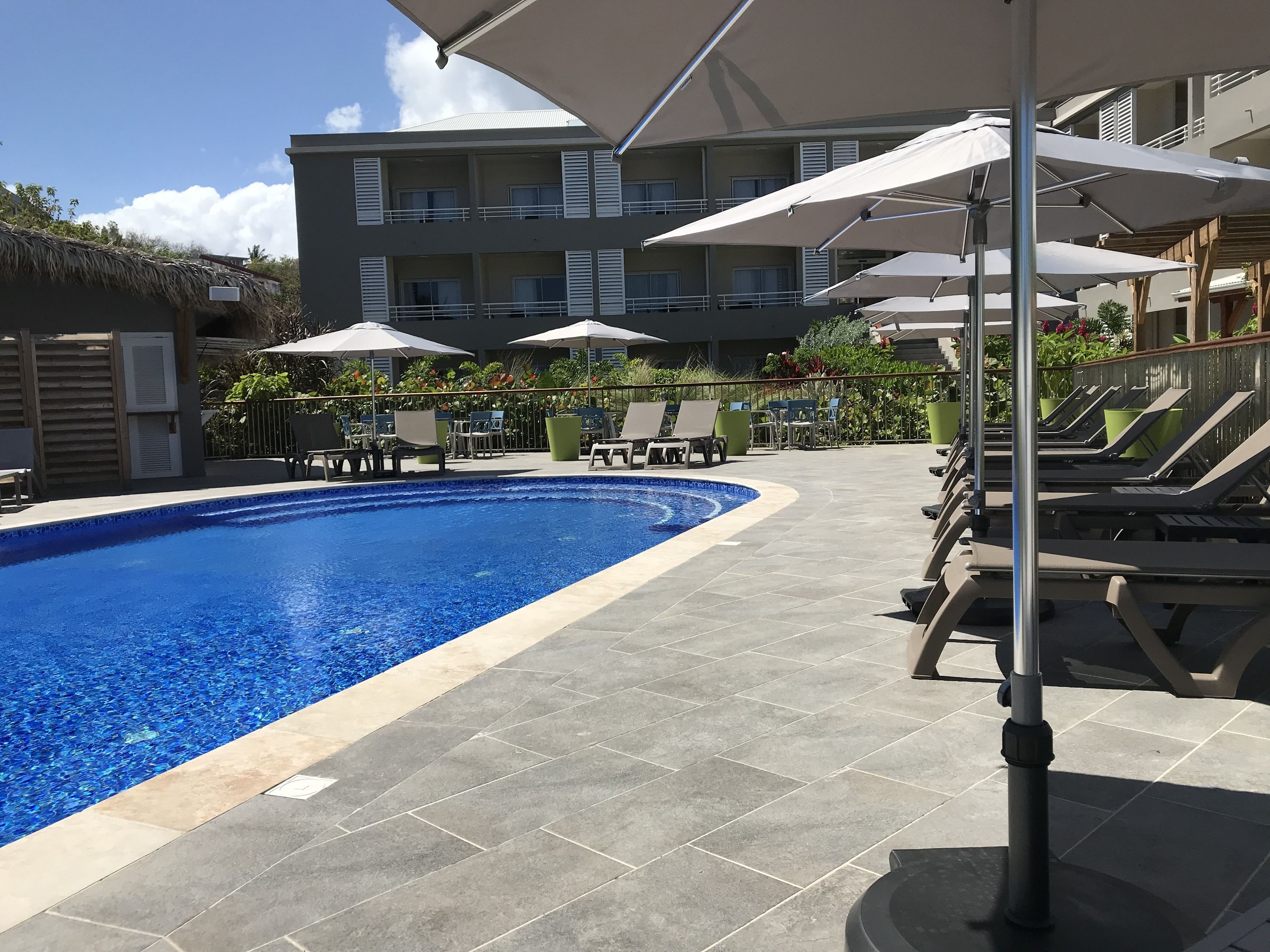 A swimming pool with lounge chairs and umbrellas, surrounded by a building with balconies and a thatched roof structure.