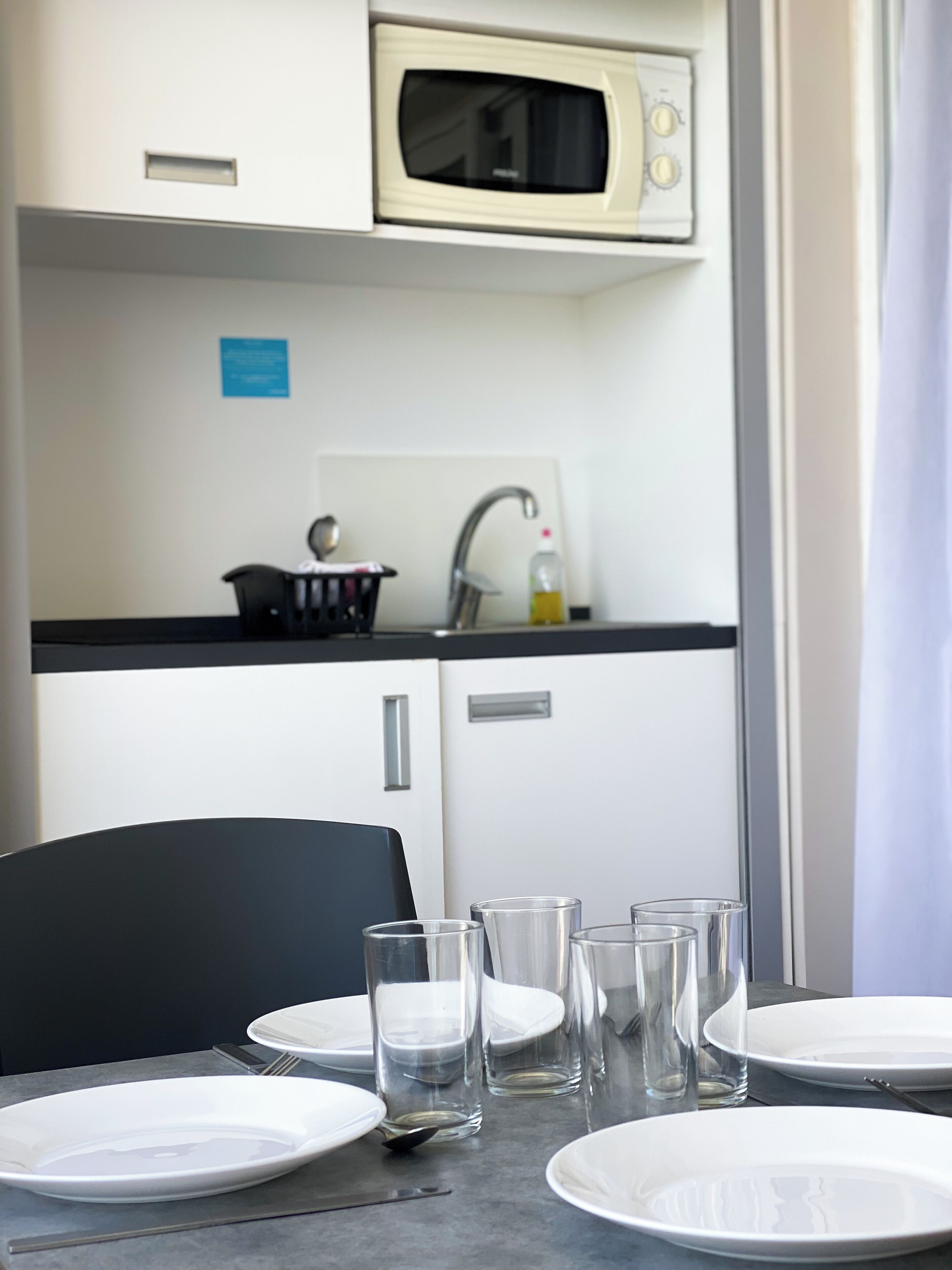 Une petite cuisine équipée d’un four à micro-ondes, d’un évier et d’une petite chaise noire. Il y a des assiettes et des verres vides sur la table.