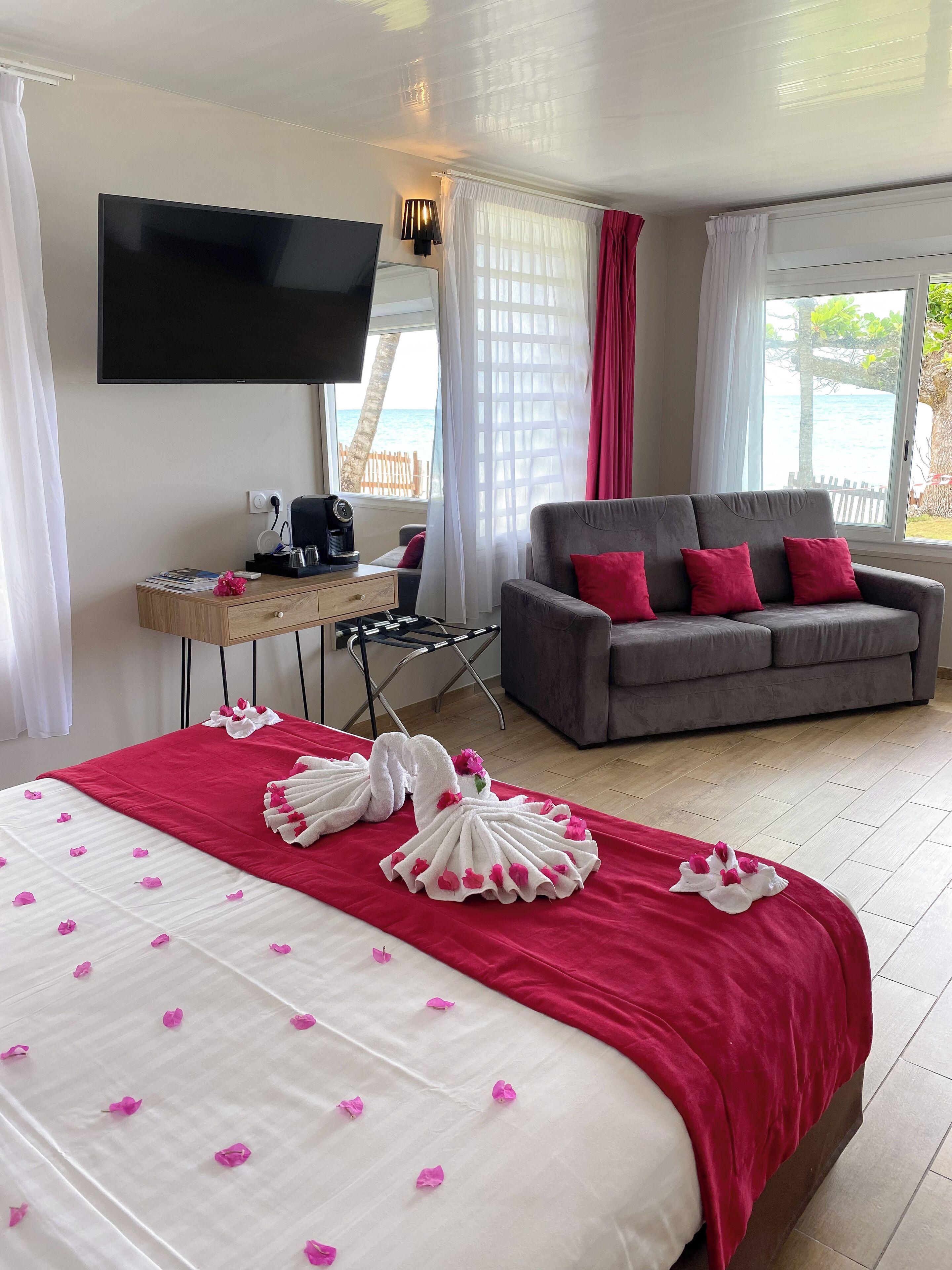A hotel room with a bed, a grey sofa, a TV, and a window with a view of the sea.