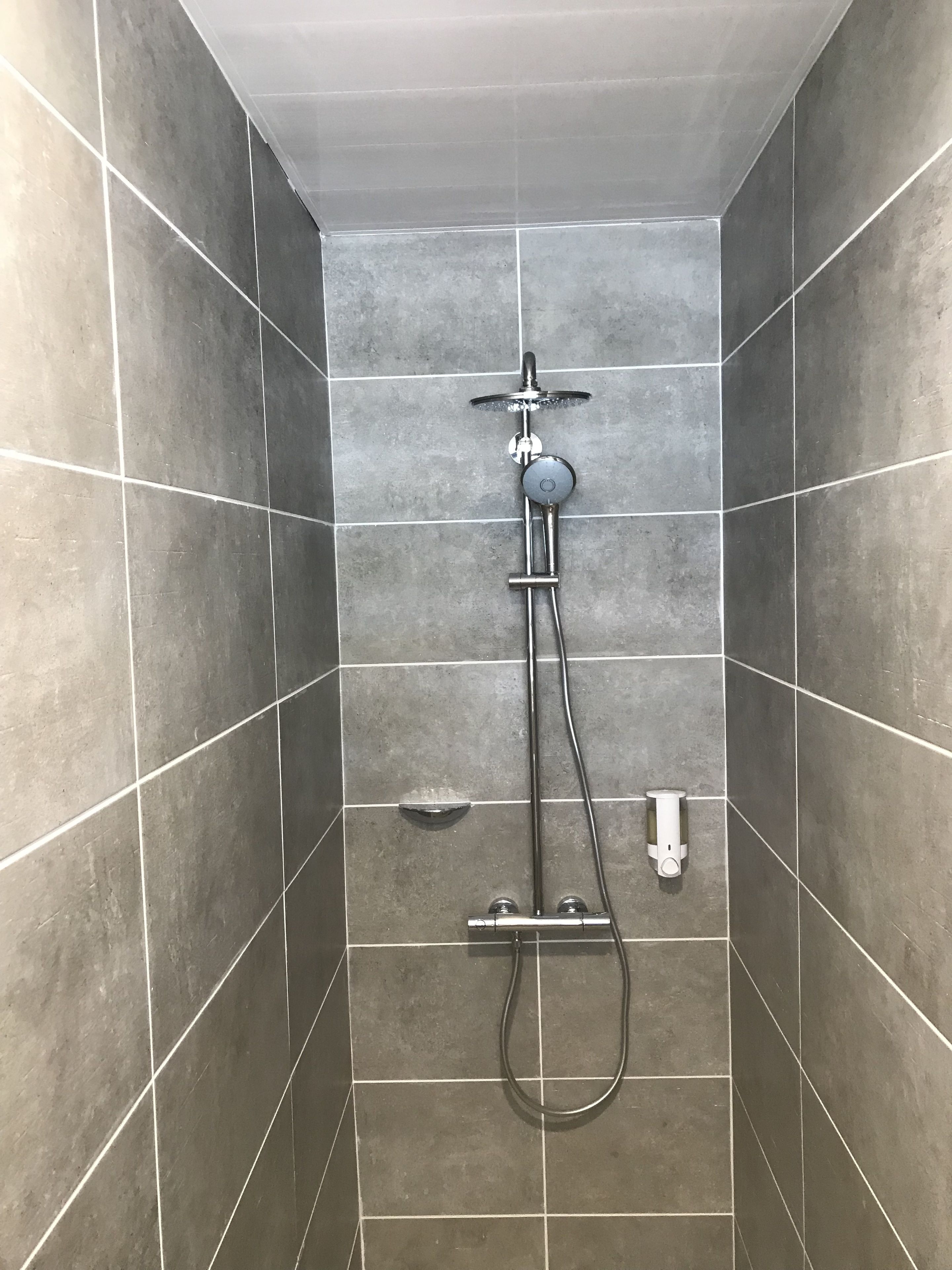 A shower with a handheld showerhead and a wall-mounted faucet, set in a tiled room.