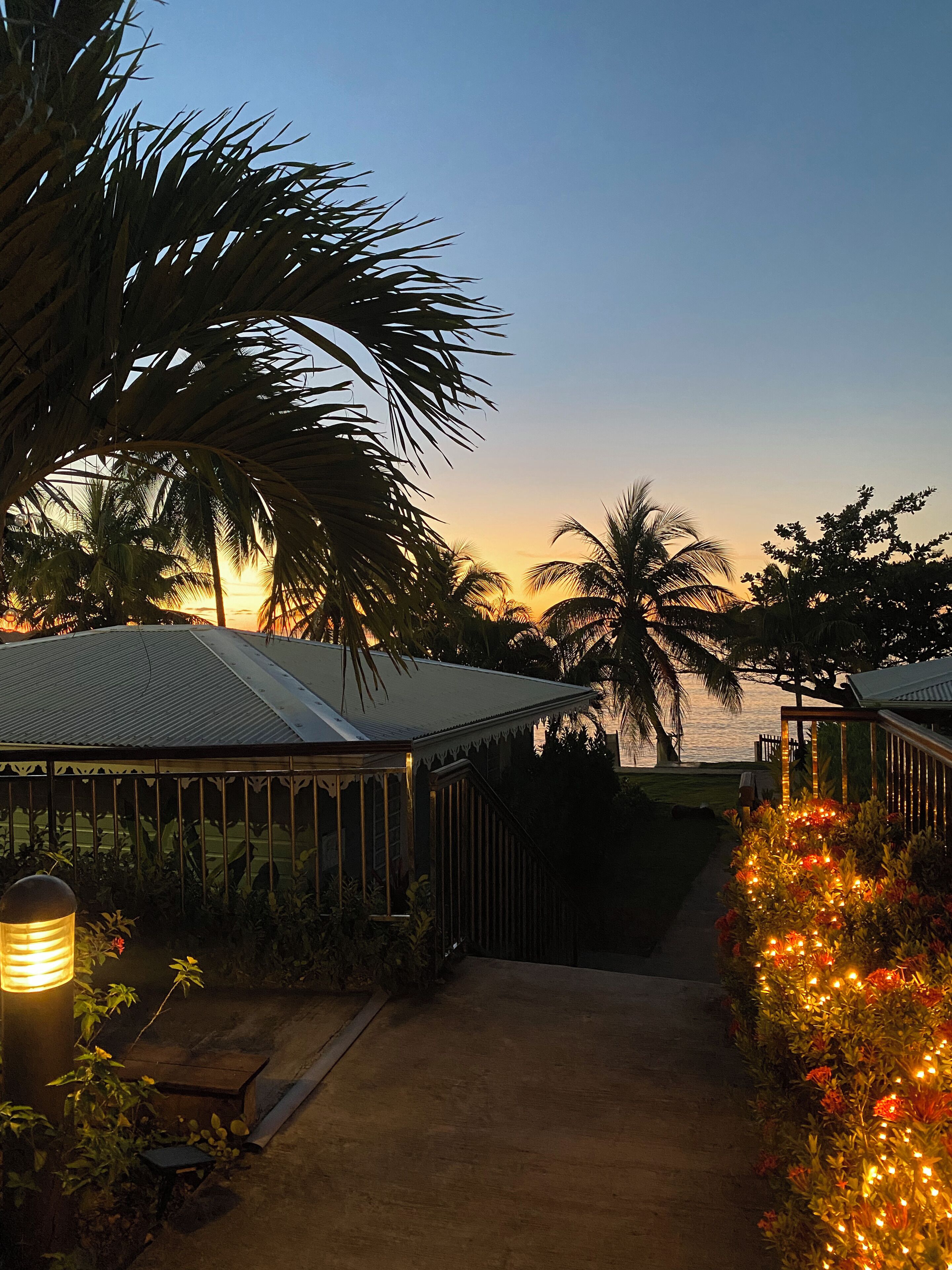 Un sentier mène à une zone abritée d’où l’on peut admirer le coucher de soleil sur l’eau, le tout étant entouré de palmiers et d’un jardin fleuri.
