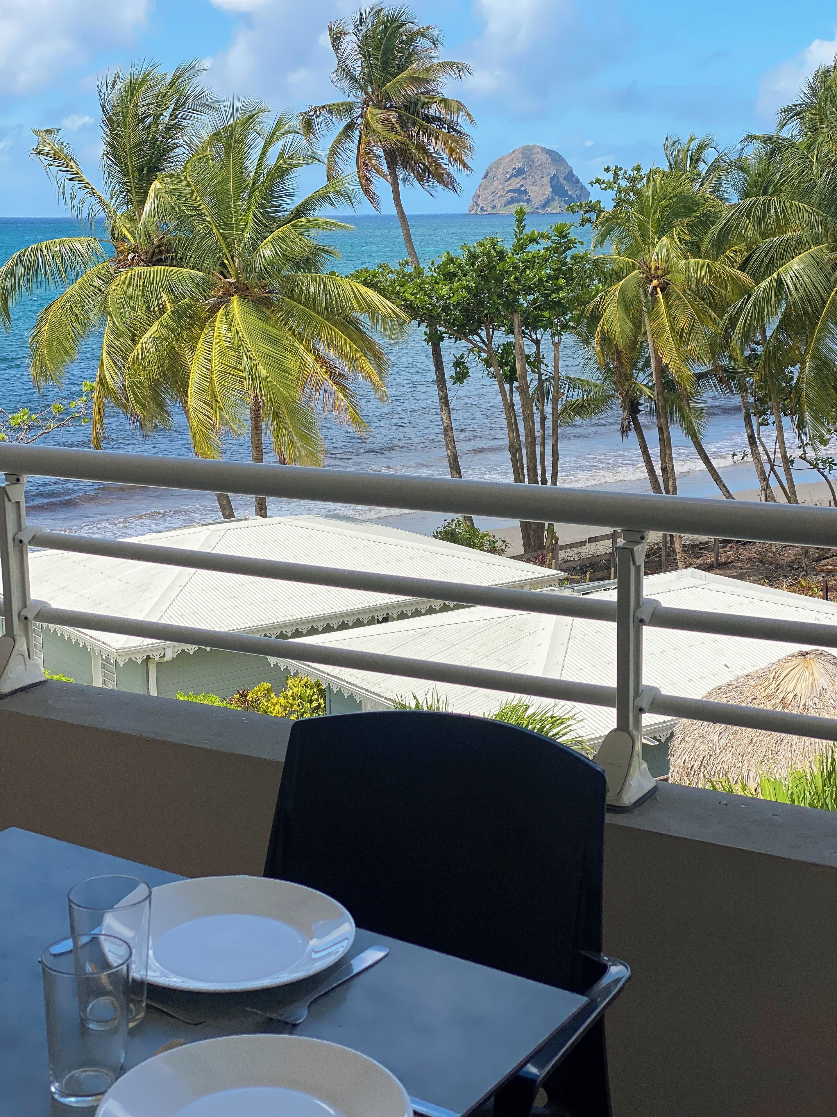 A balcony with a view of the ocean and palm trees, a table with plates and glasses, and a thatched structure in the background.