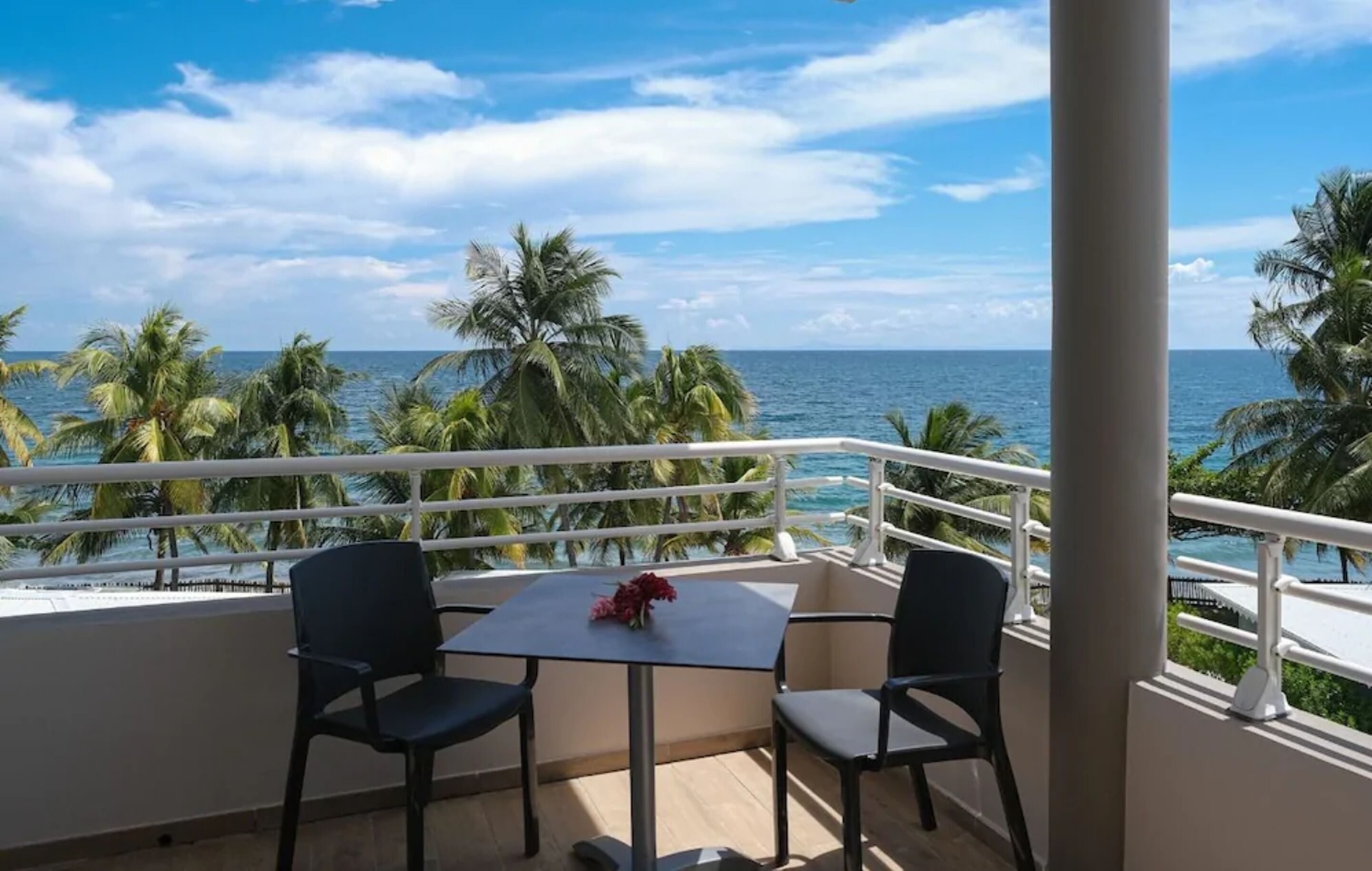 A balcony with two chairs and a table, overlooking a beach with palm trees and the ocean.