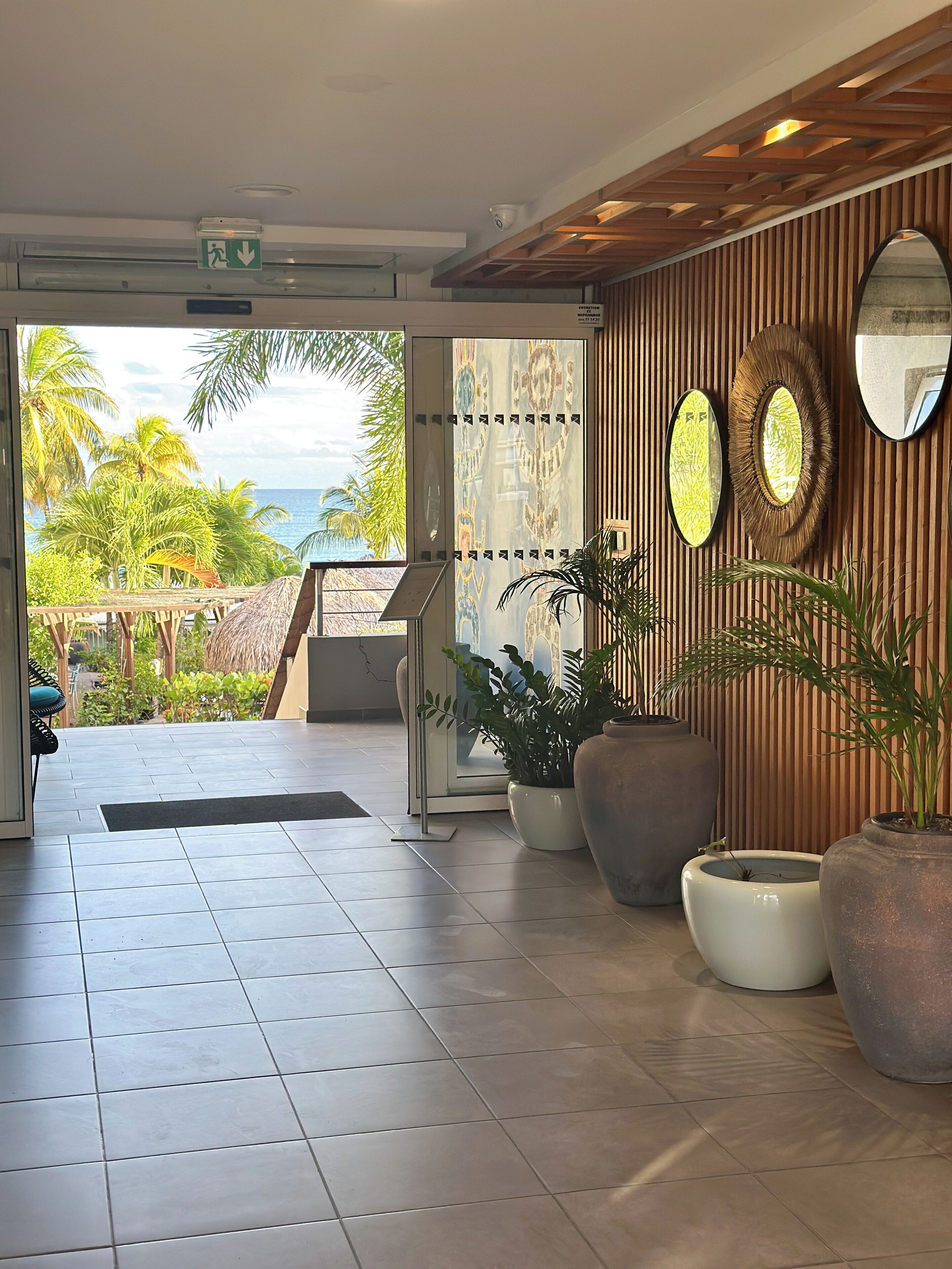 A modern hotel lobby with a view of the ocean, large potted plants, and a wooden wall with mirrors.