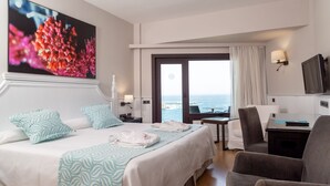 Habitación de hotel con cama, escritorio, silla, televisión y un amplio ventanal con vista al mar.