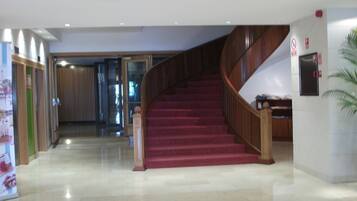 A hotel lobby with a marble floor, a wooden staircase with red carpet, and a glass door leading to another room.