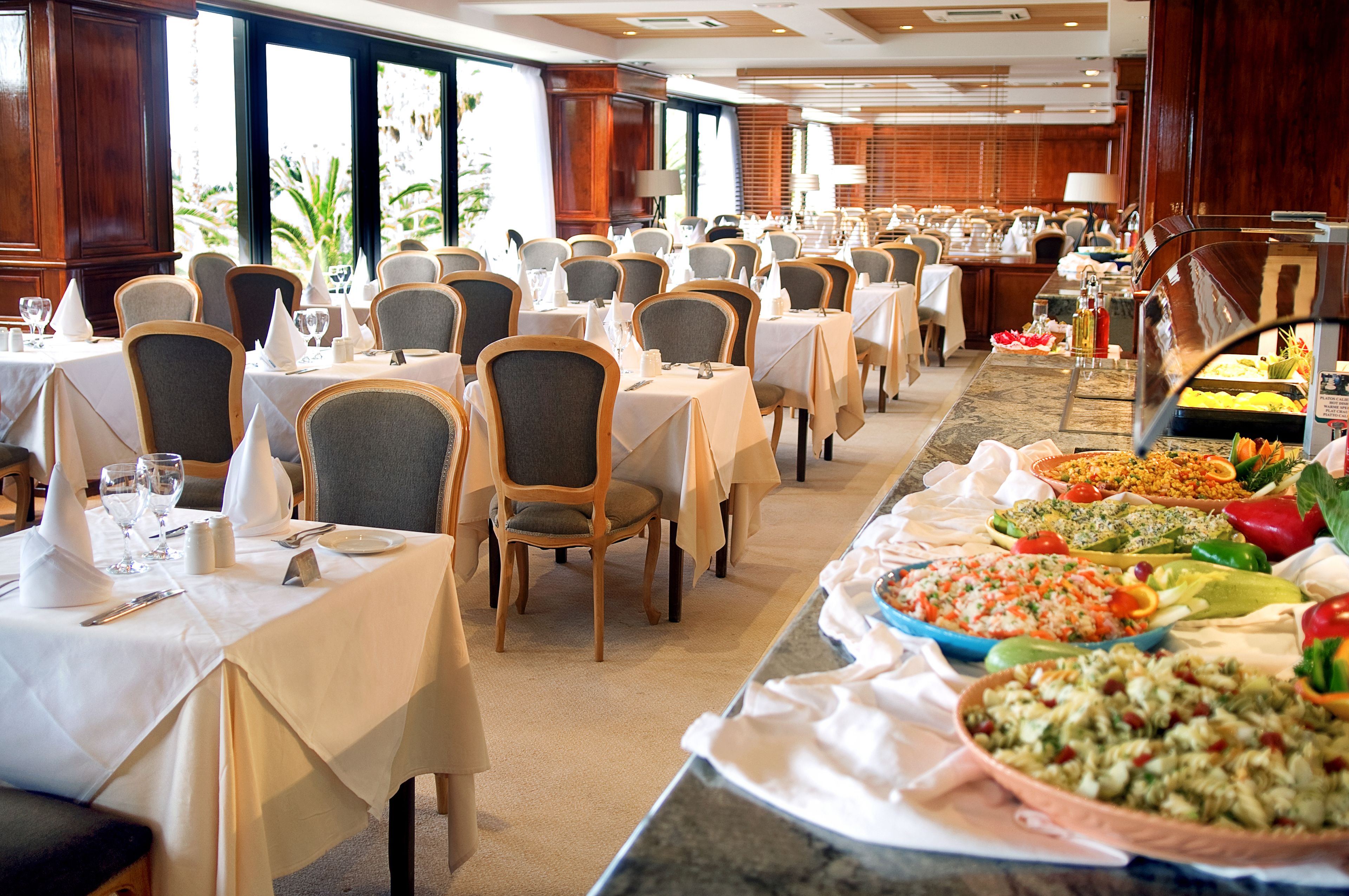 Uma mesa de buffet montada numa sala de jantar de hotel, com várias mesas e cadeiras, uma grande variedade de alimentos e vista para o exterior através de grandes janelas.