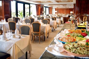 Disposición de un bufé en el comedor de un hotel, con varias mesas y sillas, una amplia variedad de alimentos y vistas al exterior a través de amplios ventanales.