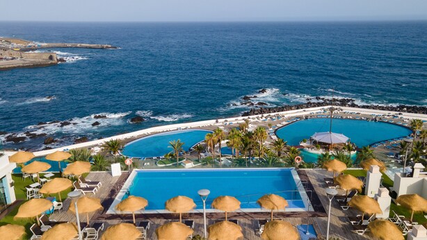 Un resort con una piscina grande, otra más pequeña y una zona de playa con sombrillas.