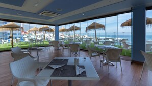 Un restaurante con mesas y sillas blancas, amplias ventanas con vistas al océano y sombrillas de paja.