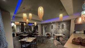 A modern restaurant with high ceilings, hanging woven light fixtures, and a variety of seating arrangements including tables and bar stools. - Roots Hotel Apartment (Accra)