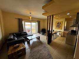 A modern living room with a sofa, coffee table, and a dining area with a table and chairs. There is a kitchen area with a stove and a painting on the wall.