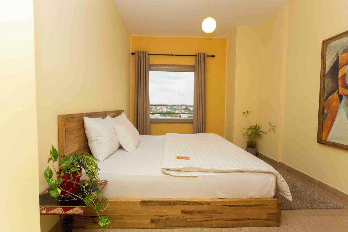 A bedroom with a wooden bed, white bedding, a window with curtains, a plant on the nightstand, and a painting on the wall.