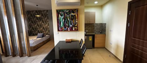 A modern living room with a glass dining table, a sofa, and a kitchen area with wooden cabinets and a built-in oven.