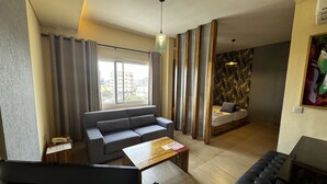 A modern living room with a grey sofa, wooden coffee table, and a black leather ottoman. A large window with curtains provides a view of the cityscape. - Roots Hotel Apartment (Accra)