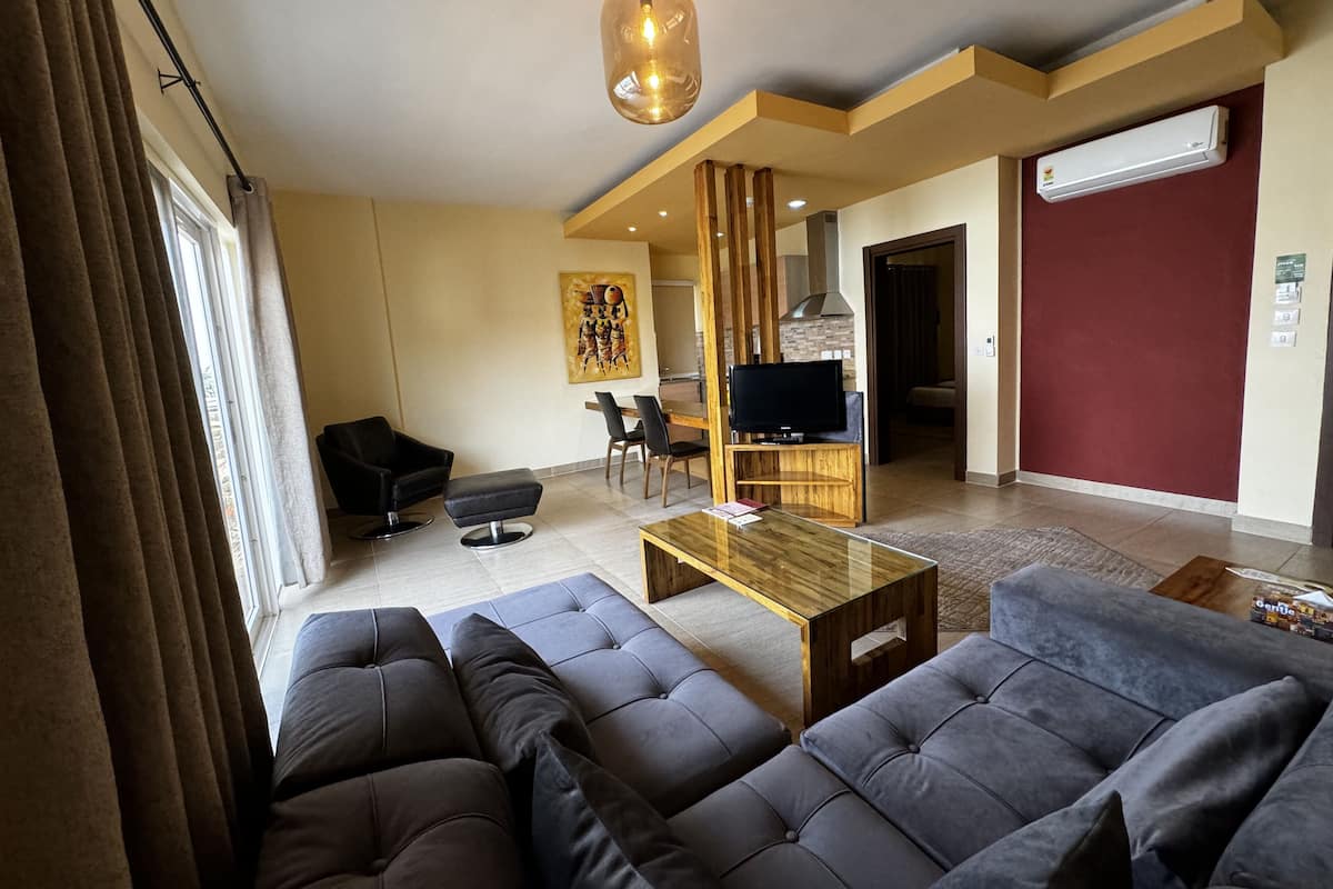 A modern living room with a sofa set, a coffee table, and a TV. There is a dining area with chairs and a table, and a kitchen in the background.