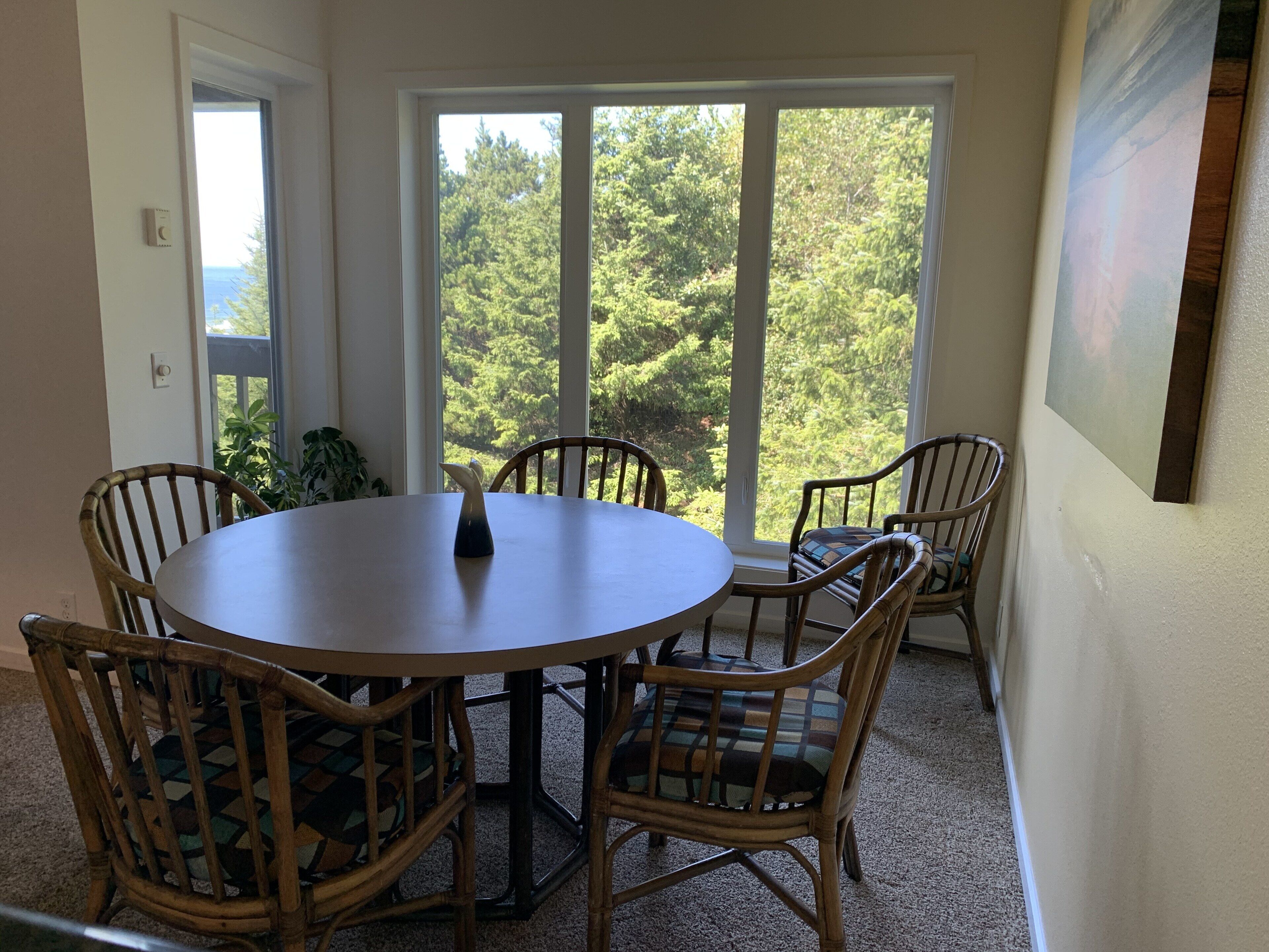 A dining area with a round table and four wicker chairs, large windows with a view of trees, and a painting on the wall.