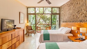 Habitación de hotel con dos camas, un televisor, un escritorio con lámpara y vistas a árboles a través de ventanas amplias.