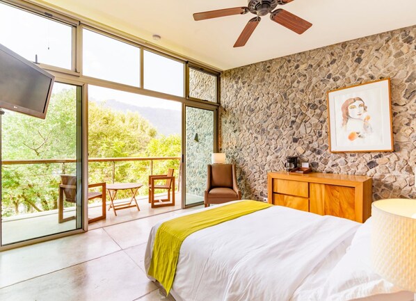 A bedroom with a large bed, a stone wall, a TV, a balcony with chairs, and a view of greenery. - Kawilal Hotel (Amatitlan)