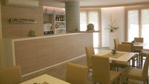 A modern dining area with a central bar, beige chairs, and a large window providing natural light. - Apart Hotel Atlantic (Grado)
