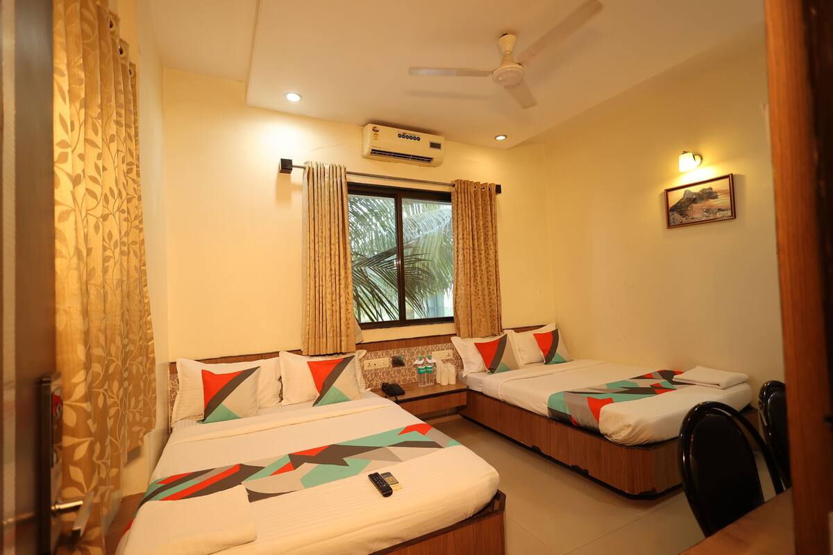 A hotel room with two beds, a ceiling fan, an air conditioning unit, a window with curtains, a bedside table with a phone and toiletries, and a wall-mounted picture.