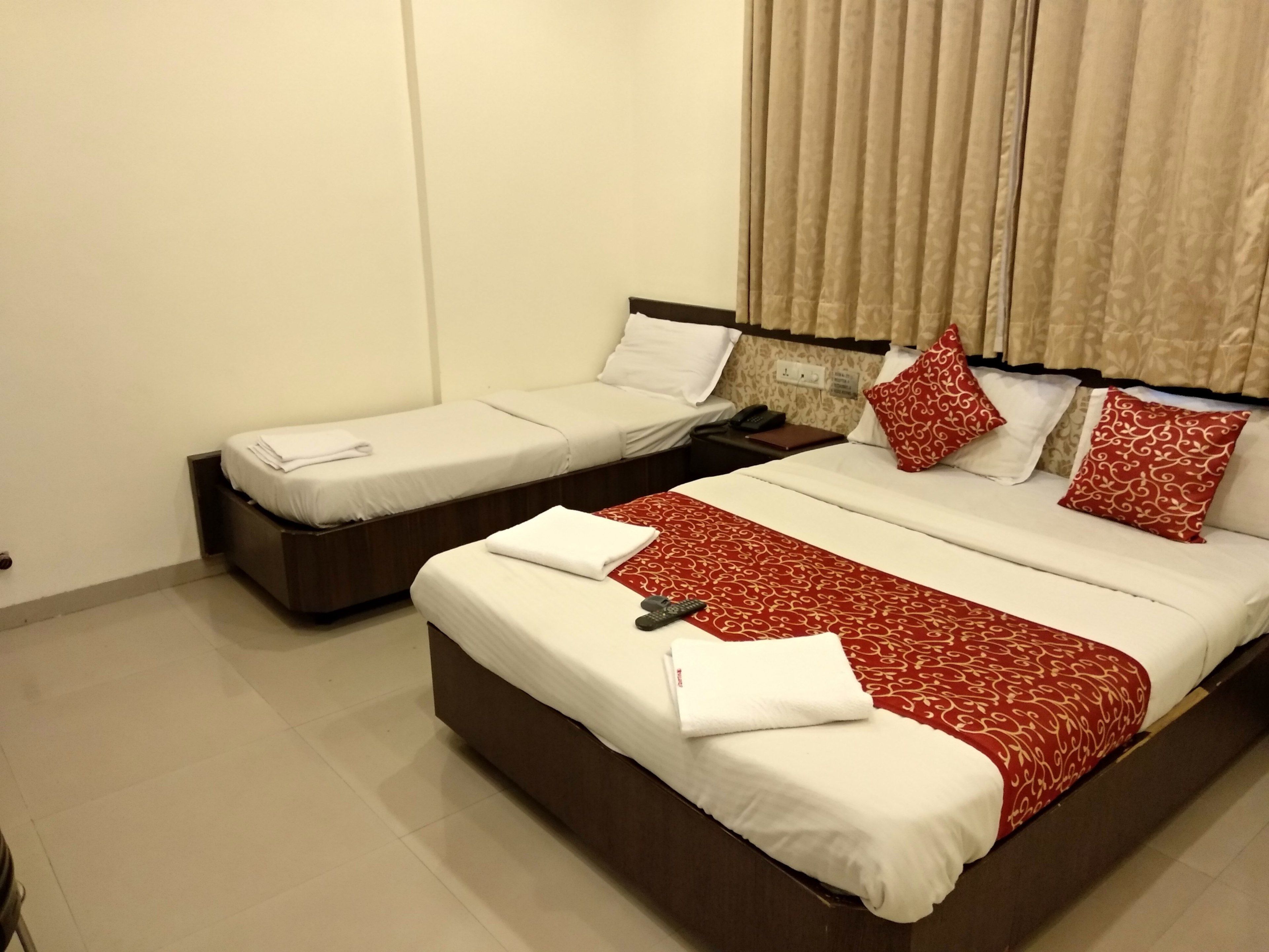 Habitación de hotel con dos camas individuales y una cama doble. Cada cama tiene sábanas blancas y colchas estampadas de rojo. Junto a la cama doble hay una mesita de noche con un teléfono y un libro. El piso es de baldosas y las cortinas de la ventana son color beige.