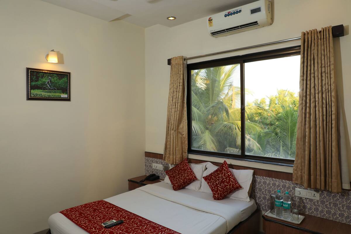 A hotel room with a large window, a bed with white bedding and red pillows, a bedside table with a phone and a glass of water, and an air conditioning unit on the wall.