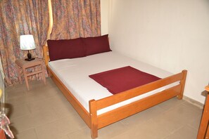A single bed with a wooden frame, a small bedside table with a lamp, and a floral curtain. - Pink Hostel (Accra)
