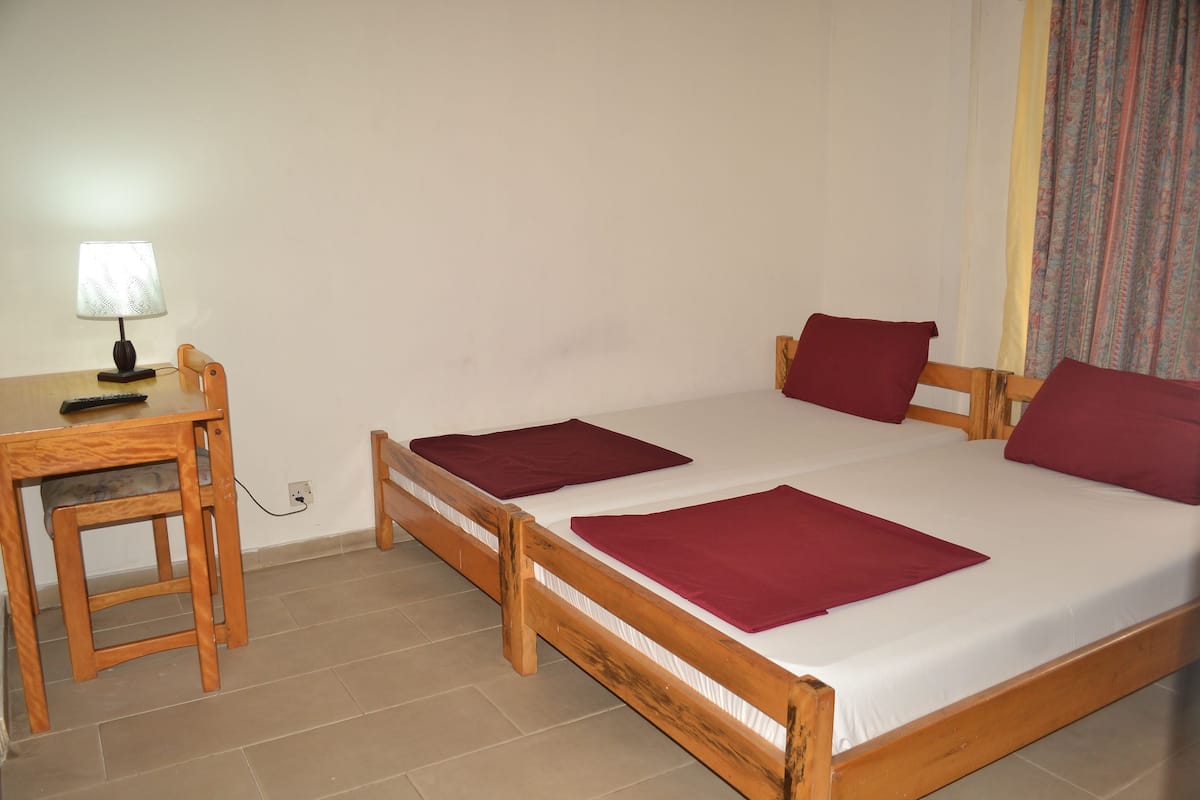 Quarto simples com duas camas de madeira, uma escrivaninha com abajur e uma cadeira.