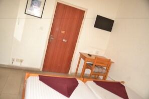 Ein Hotelzimmer mit Holztür, einem an der Wand befestigten Fernseher, einem kleinen Holztisch und zwei Betten mit violetten Überwürfen.