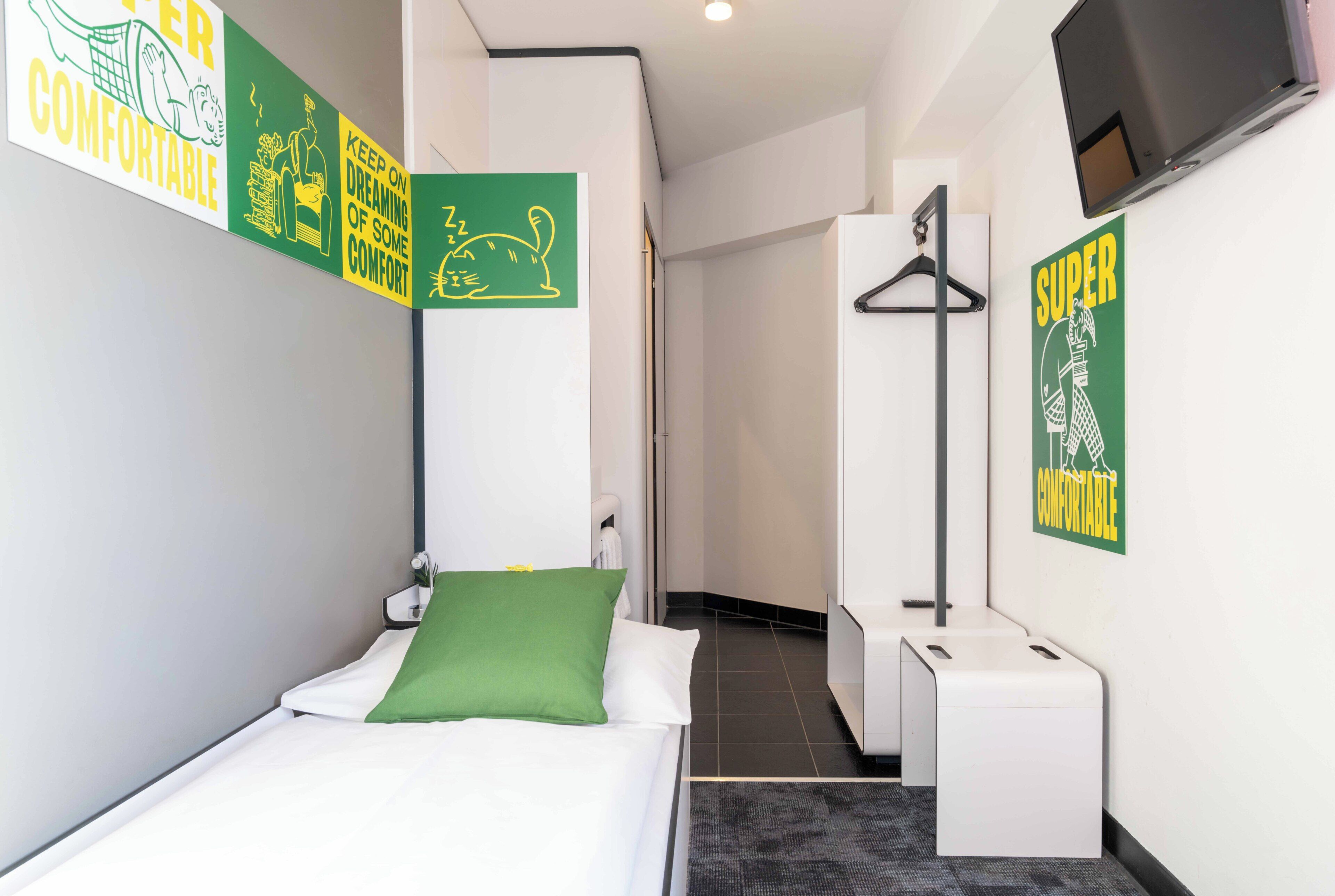 Una habitación de hotel con una cama, un almohadón verde, una pared blanca, un televisor y un cartel que dice 'SUPER COMFORTABLE'.