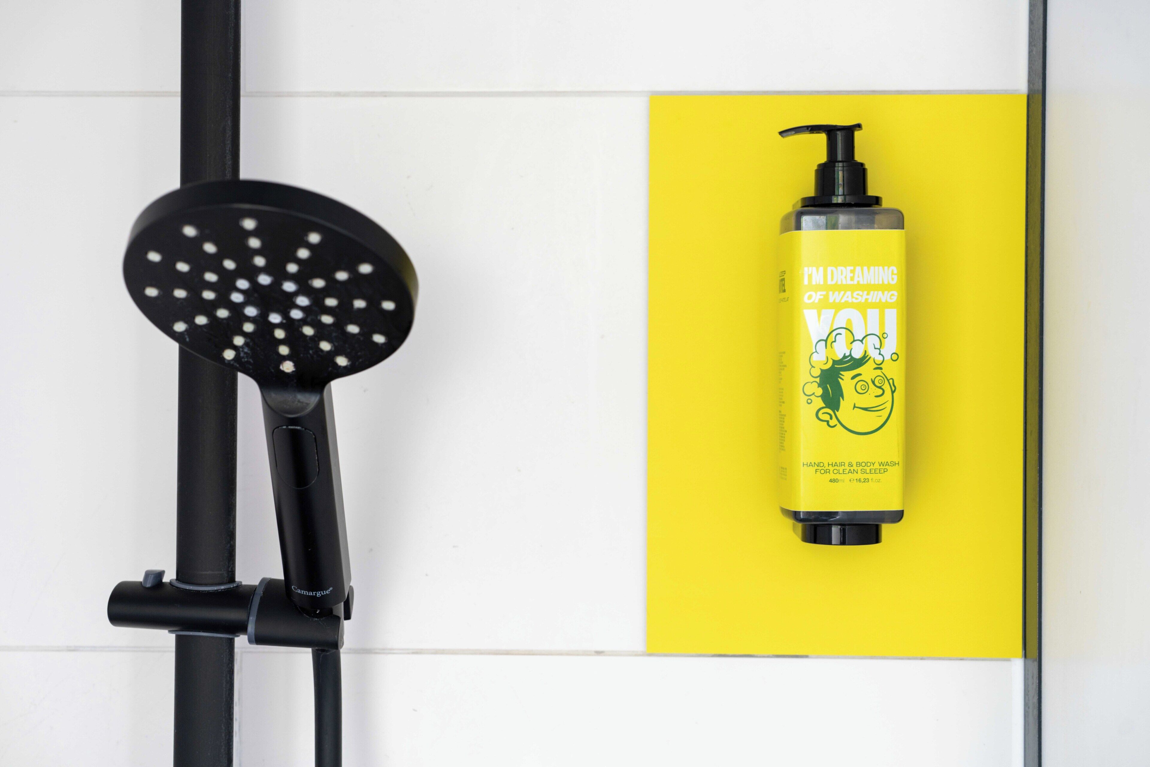 Un cabezal de ducha, una botella amarilla de jabón líquido para manos, cabello y cuerpo, y una pared de azulejos blancos.