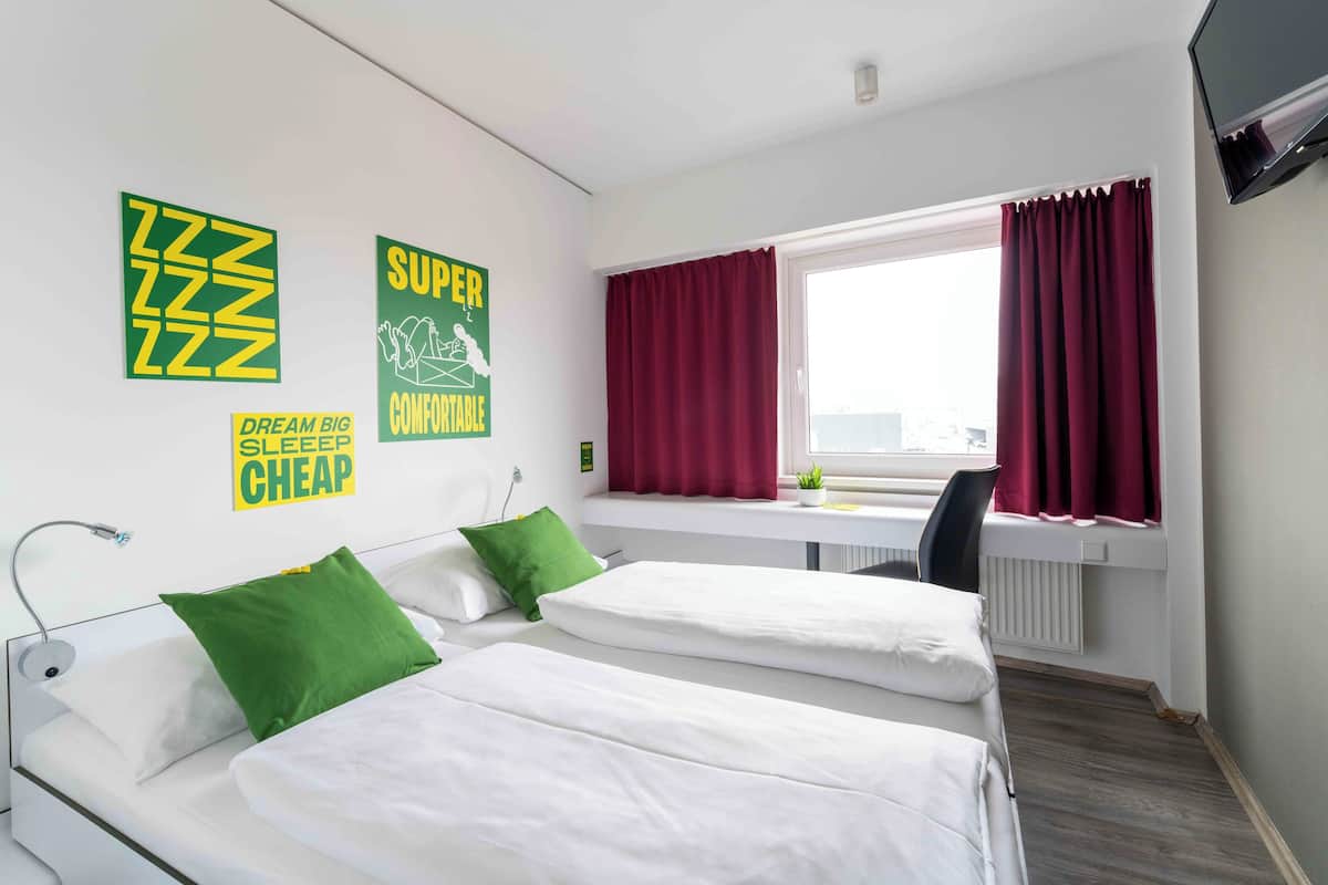 A hotel room with two beds, a window with red curtains, a desk, and a TV mounted on the wall.