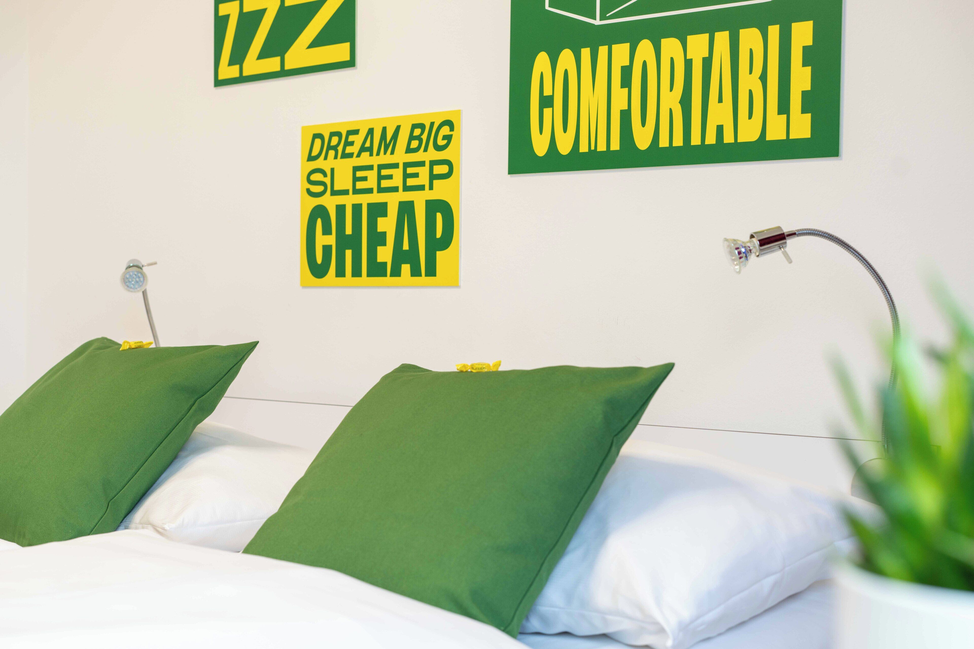 Un lit bien fait, avec des oreillers verts et blancs, une lampe fixée au mur et des affiches motivantes.