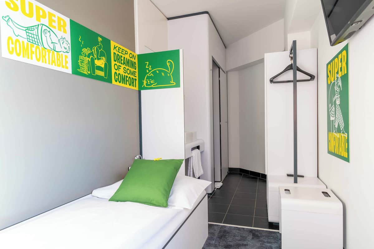 A hotel room with a bed, a green pillow, a towel rack, and a wall with signs.