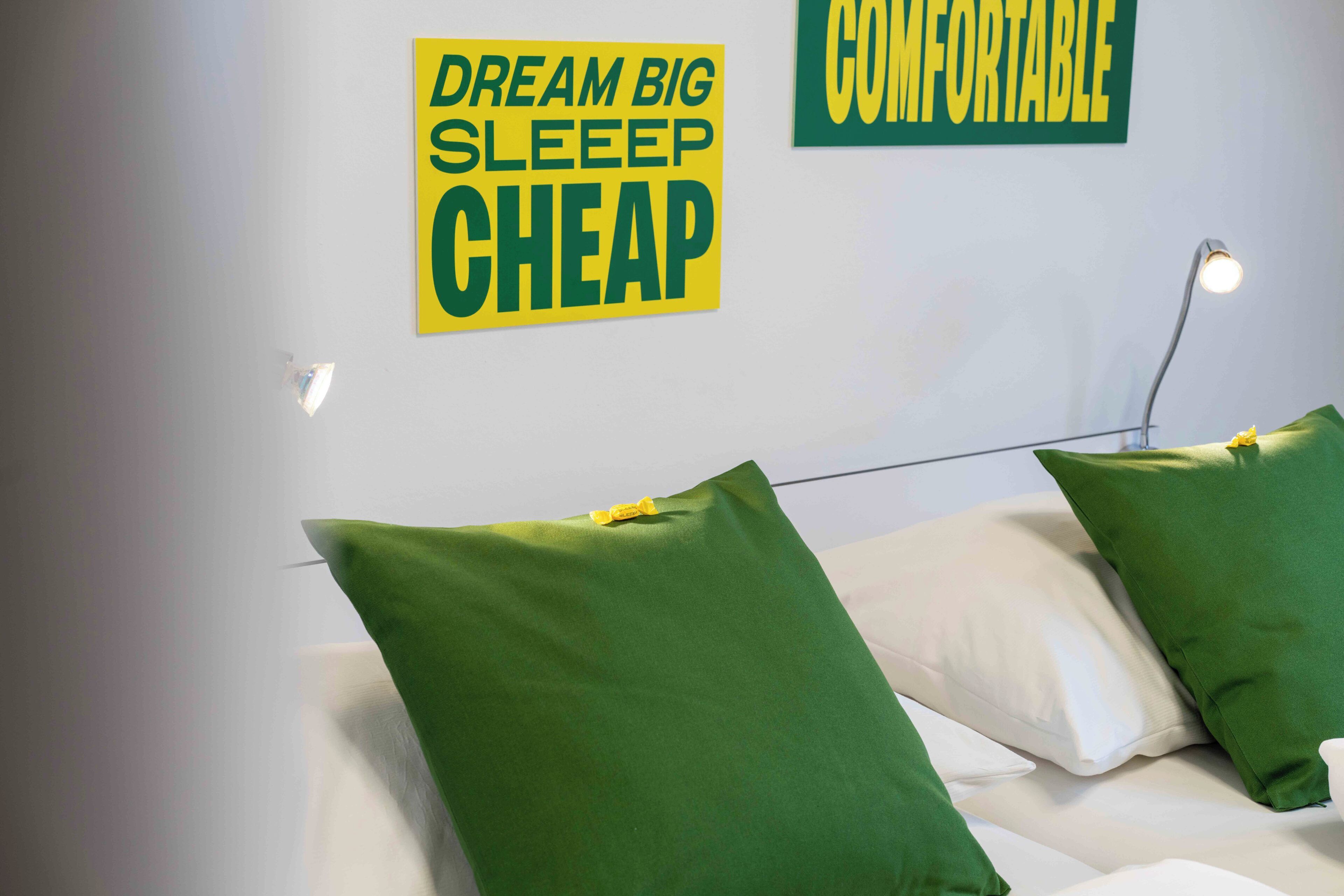 A bed with green pillows and a sign on the wall that reads "DREAM BIG SLEEP CHEAP" and "COMFORTABLE".
