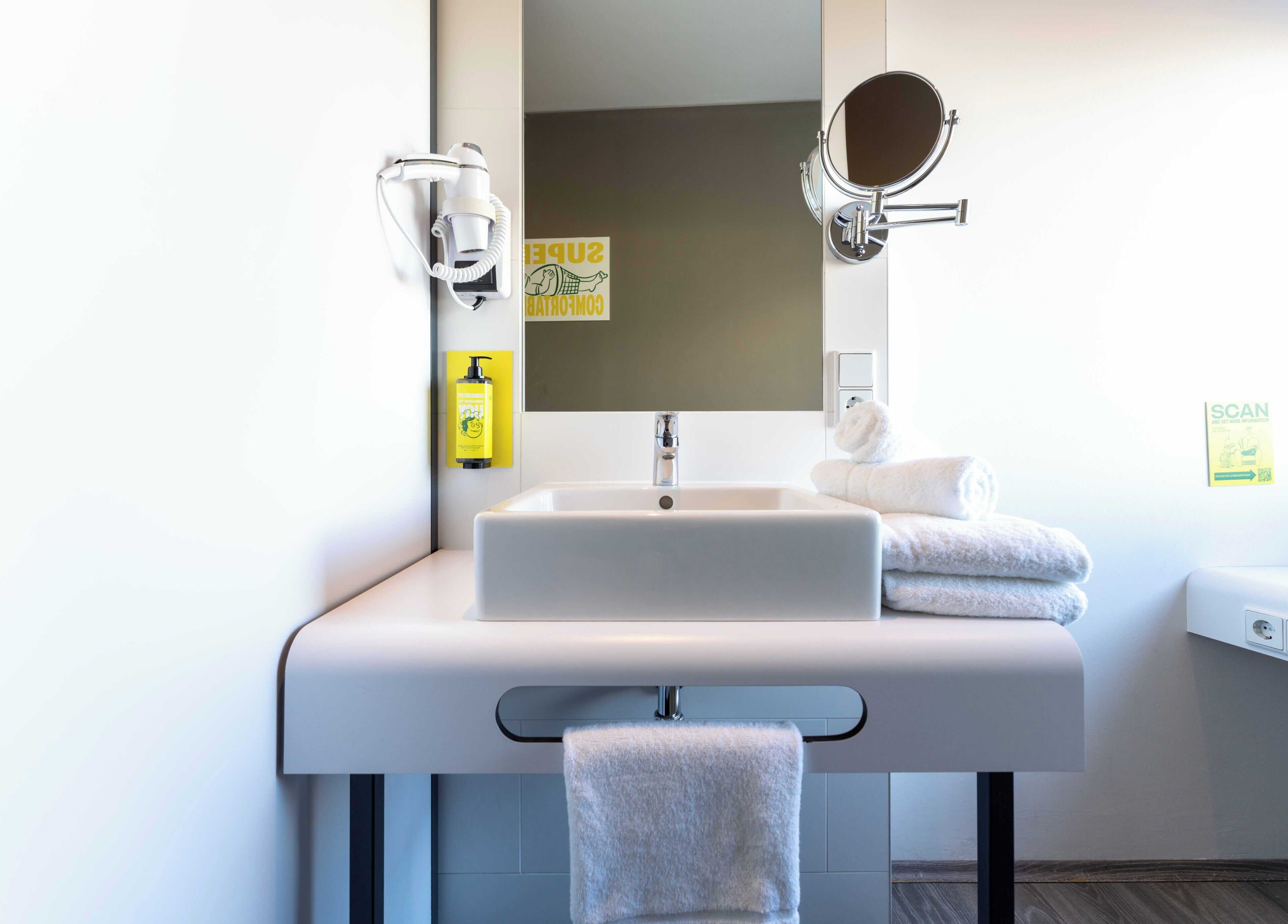 Ein modernes Badezimmer mit einem weißen Waschbecken, einem Spiegel, einem Handtuchhalter und einem Handdesinfektionsspender.