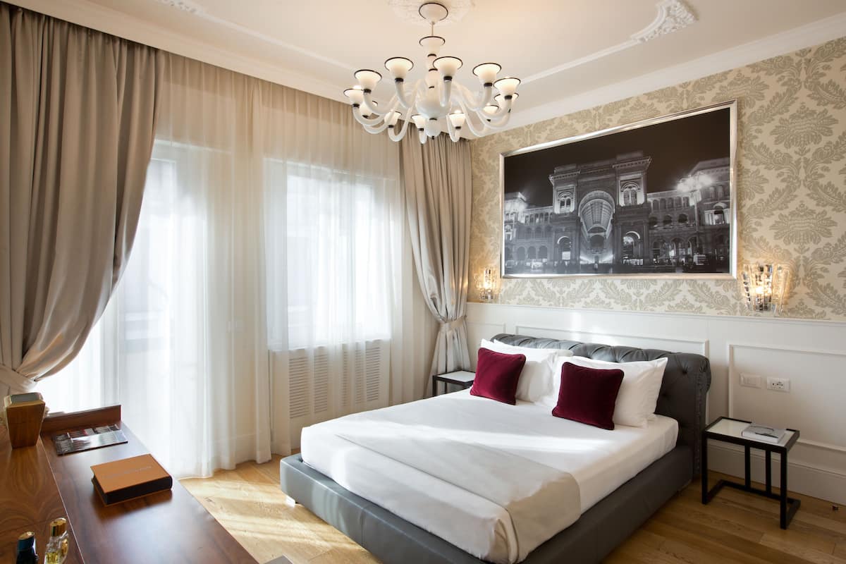 Ein Hotelzimmer mit einem großen Bett, einer Kandelaber, einem Nachttisch, einem Schreibtisch und einem großen, gerahmten Stadtbild.