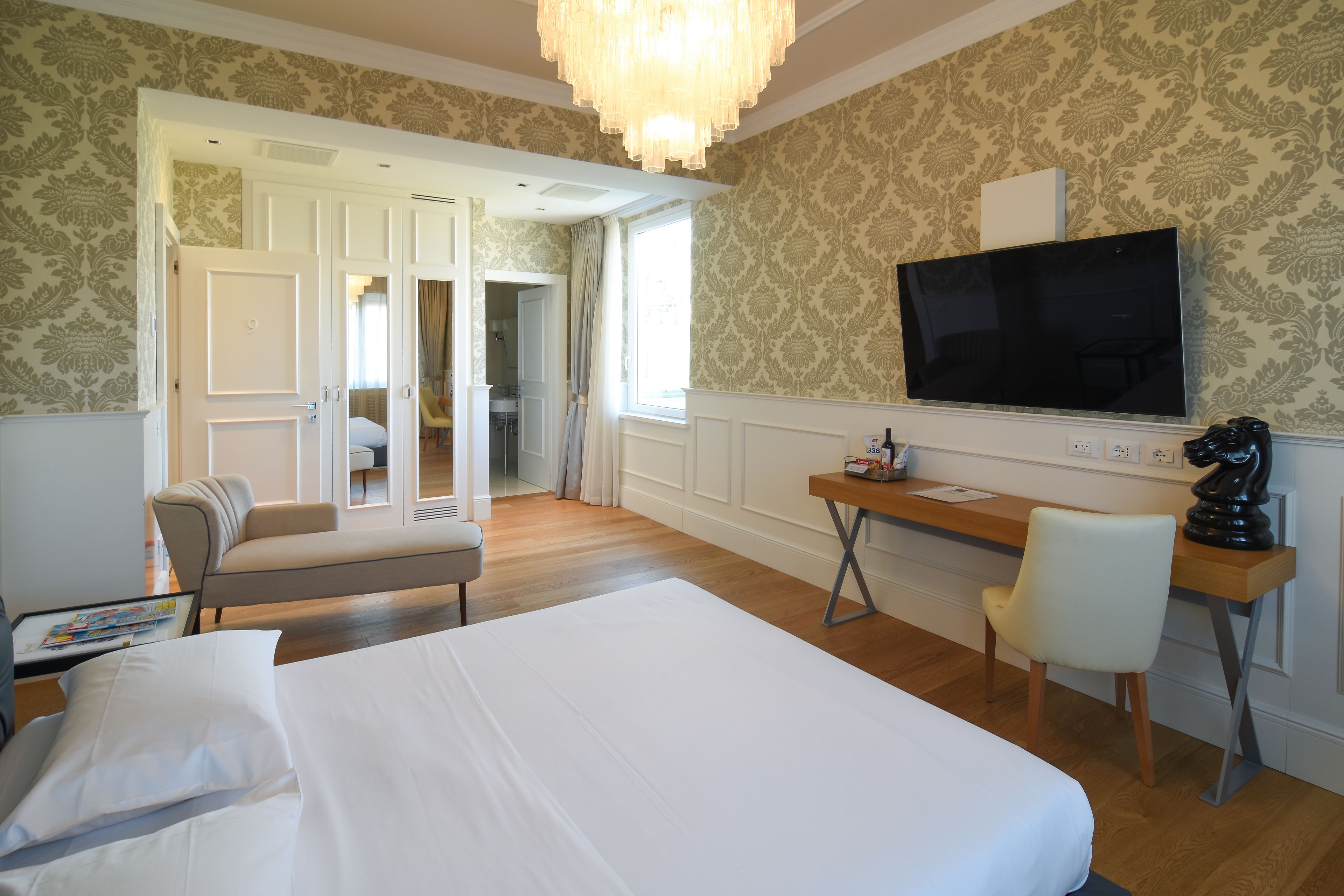 A hotel room with a large bed, a desk with a chair, a television, and a view of the hallway.