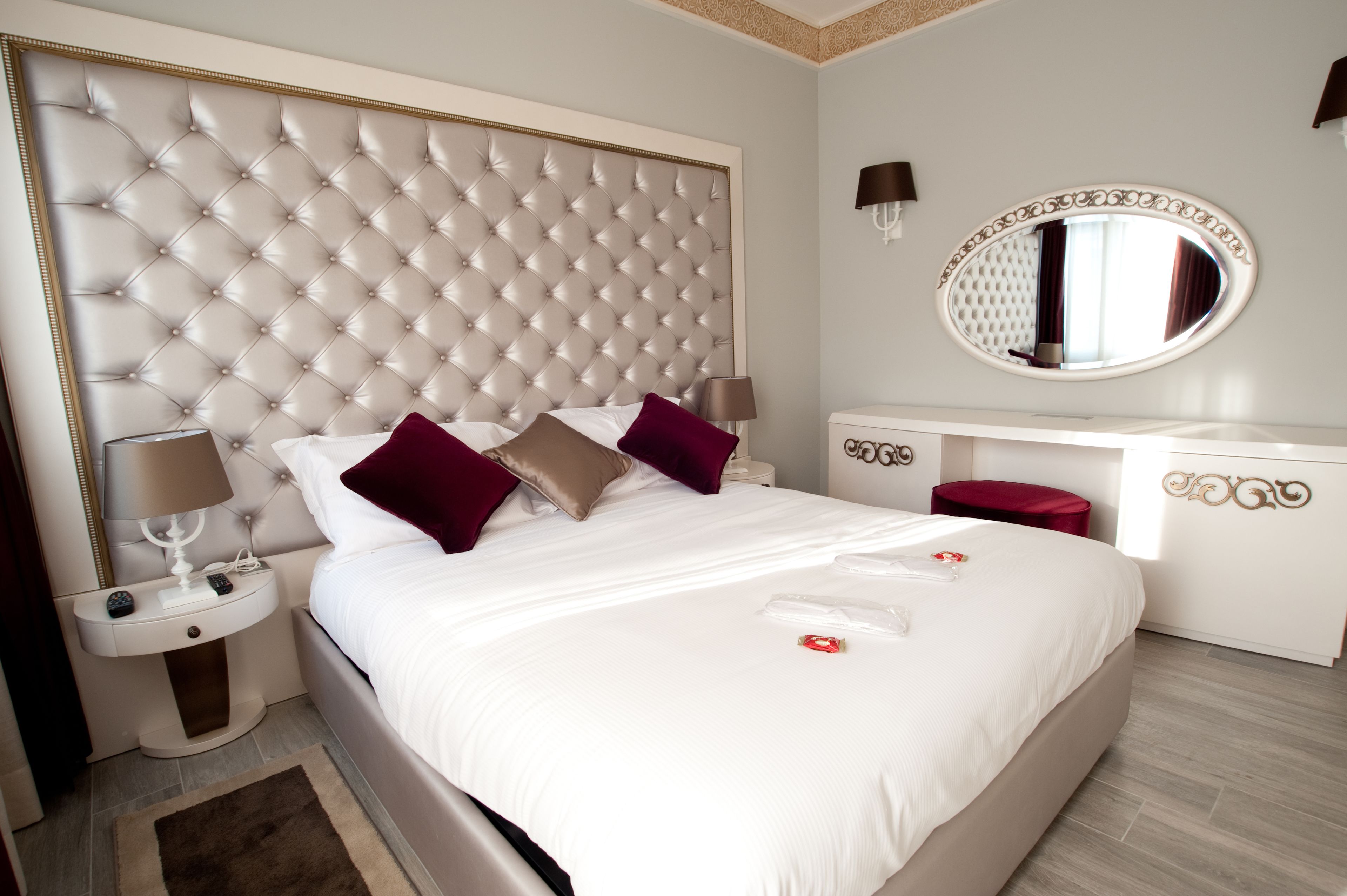 Une chambre d’hôtel avec une tête de lit capitonnée, un miroir rond, un bureau blanc et un pouf rouge.