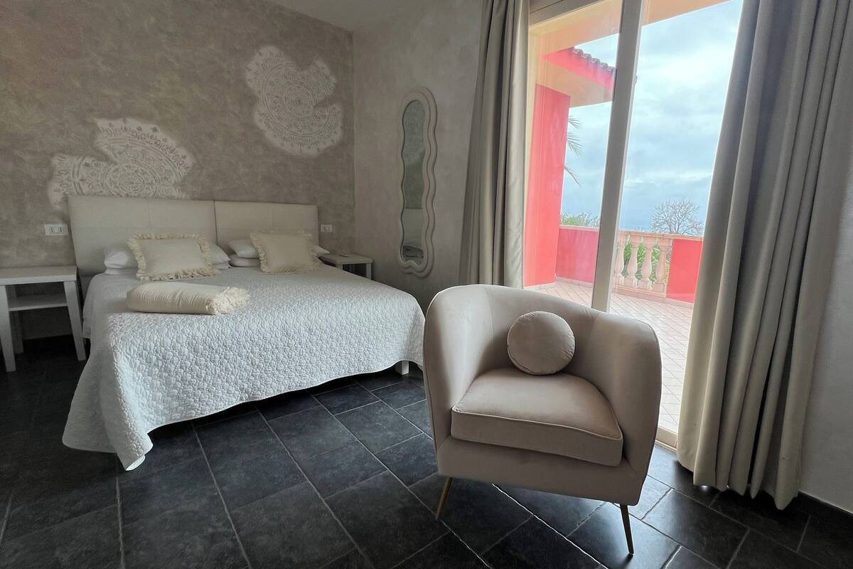 A bedroom with a bed, a nightstand, a chair, and a view of the outside.