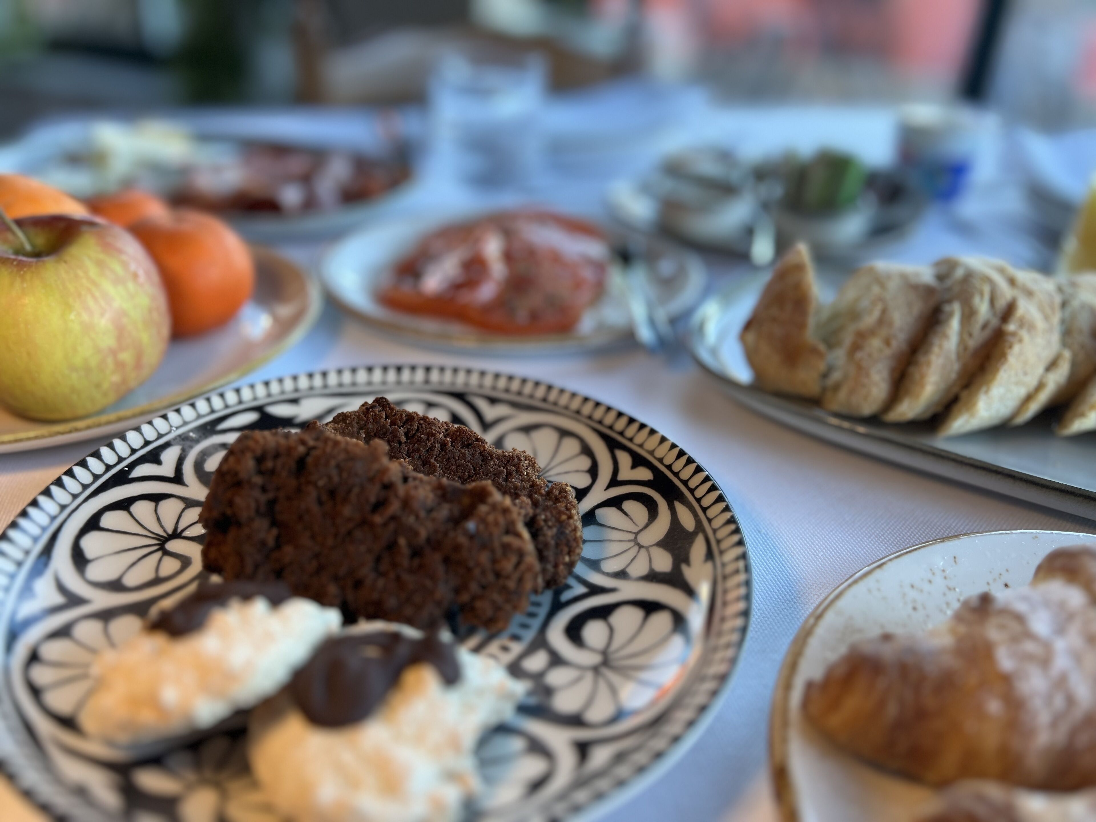 Uma mesa com diversos alimentos, incluindo um prato com biscoitos, um prato com um croissant e um prato com frutas.