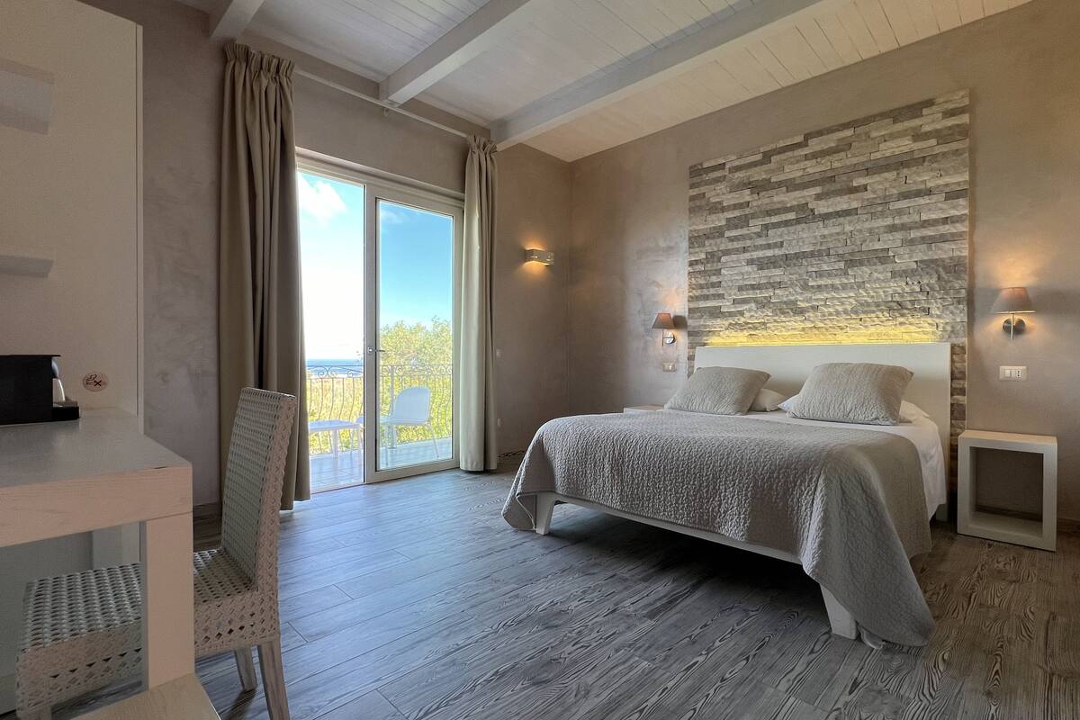 A bedroom with a large bed, a desk, and a chair. There is a balcony with a view outside.