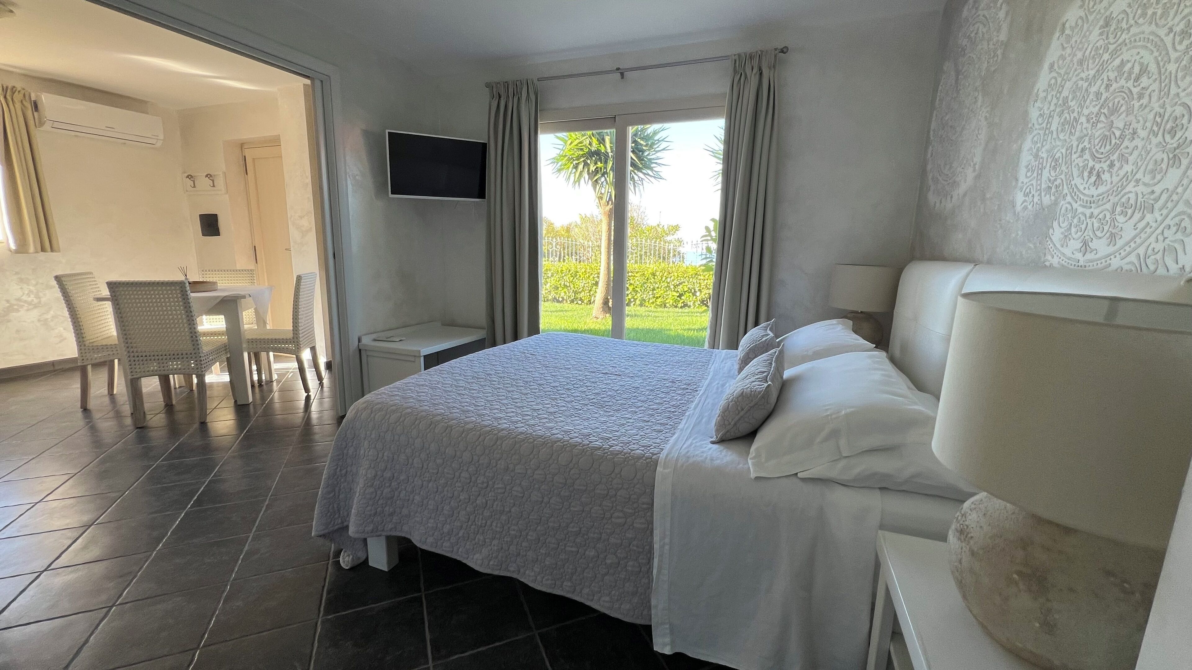 A hotel room with a bed, a TV, a desk, and a chair. There is a view of greenery outside the window.
